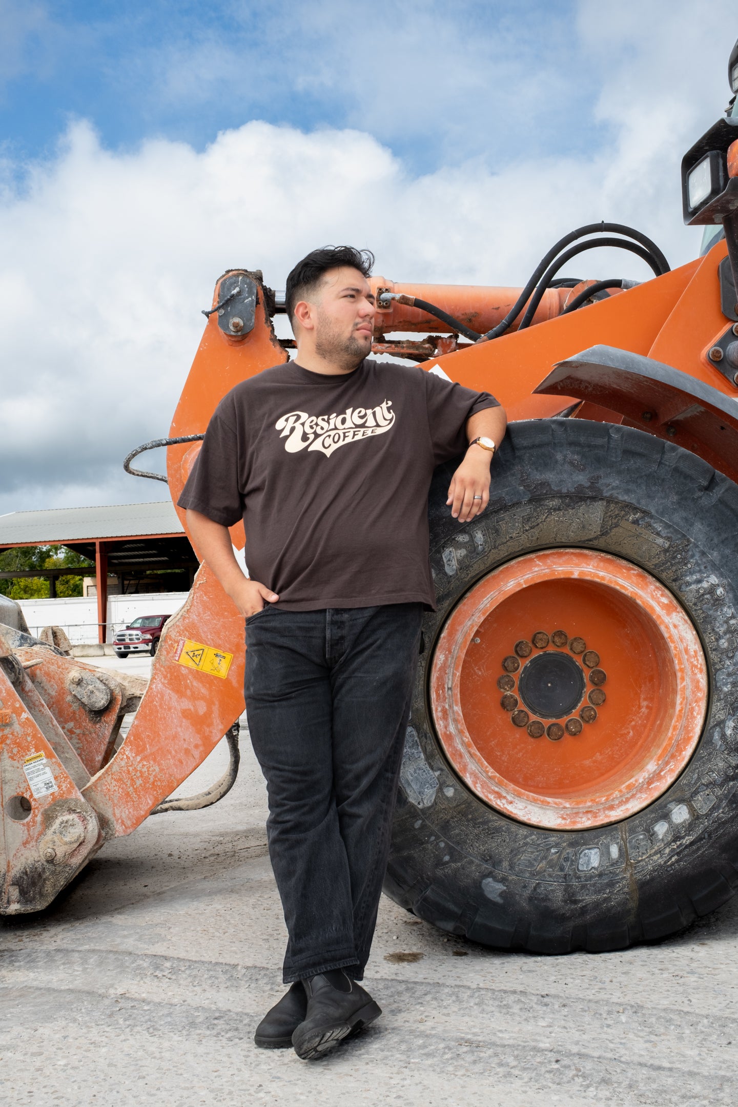 A man wearing a brown graphic t-shirt and black pants stands next to a large orange wheel loader in an outdoor setting under a blue sky.