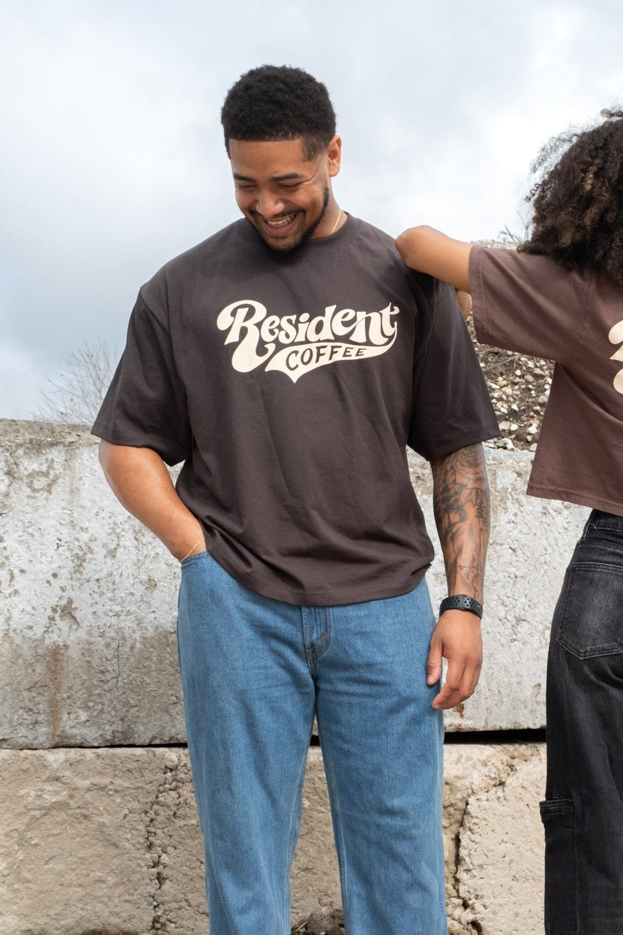Two models wearing matching brown graphic t-shirts with 'Resident Coffee' printed in white script on the chest, standing outdoors against a concrete wall under a cloudy sky.