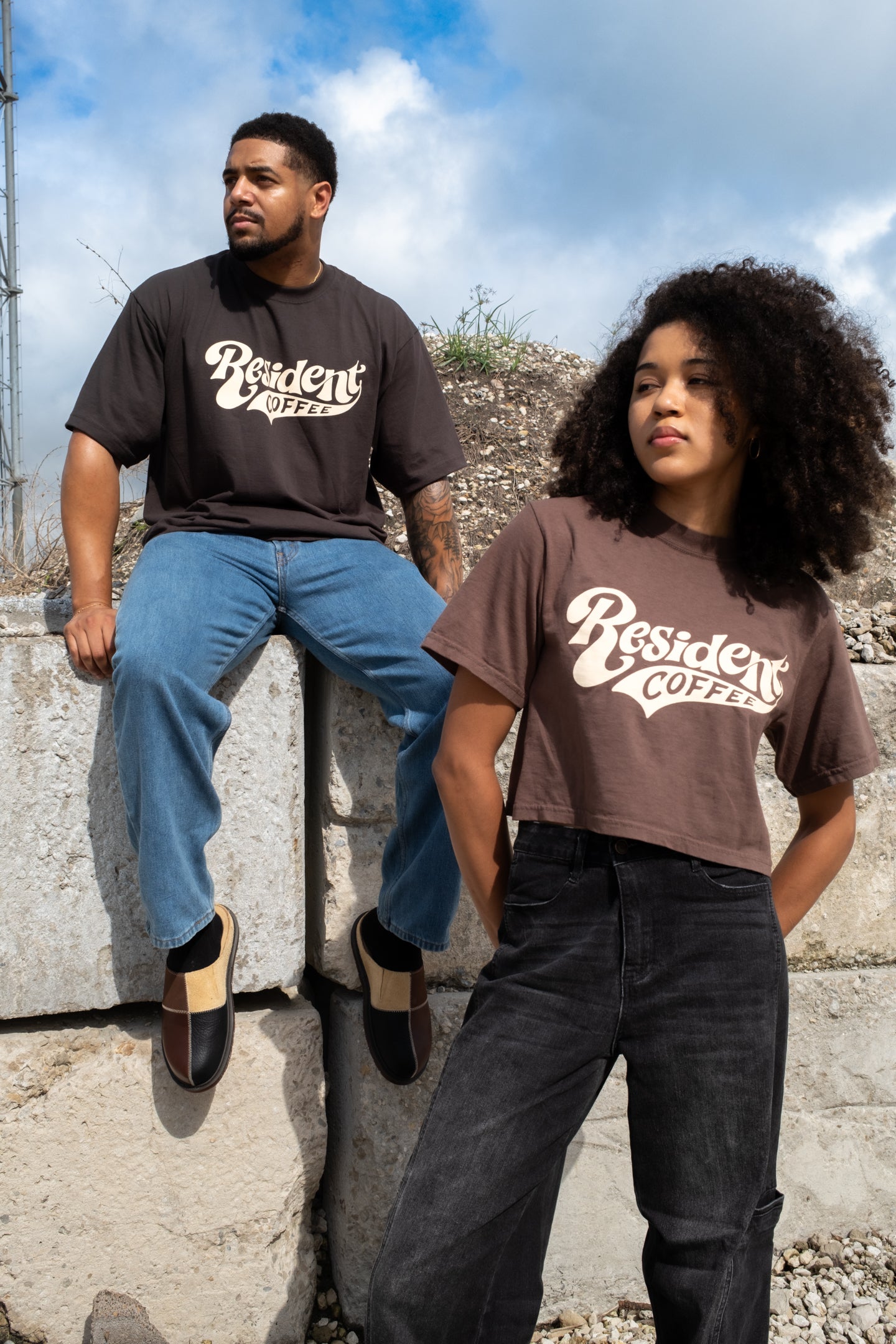 Two models wearing brown graphic t-shirts with 'Resident Coffee' printed in white script, posed outdoors against a concrete wall under a cloudy sky.