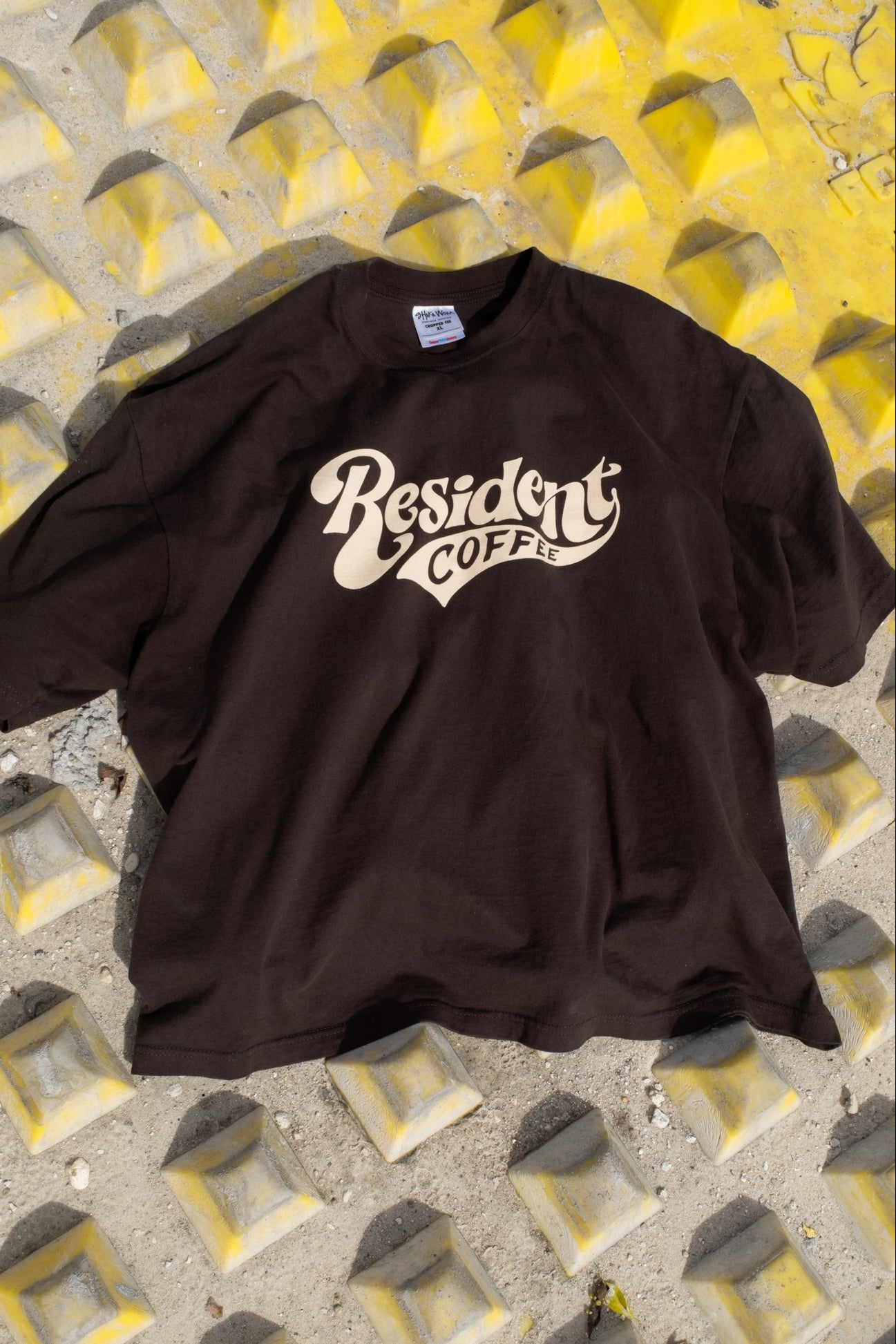 A dark brown short-sleeve t-shirt with a cream-colored 'Resident CoFFEE' graphic printed on the chest, laid flat on a textured concrete surface with yellow painted recesses.